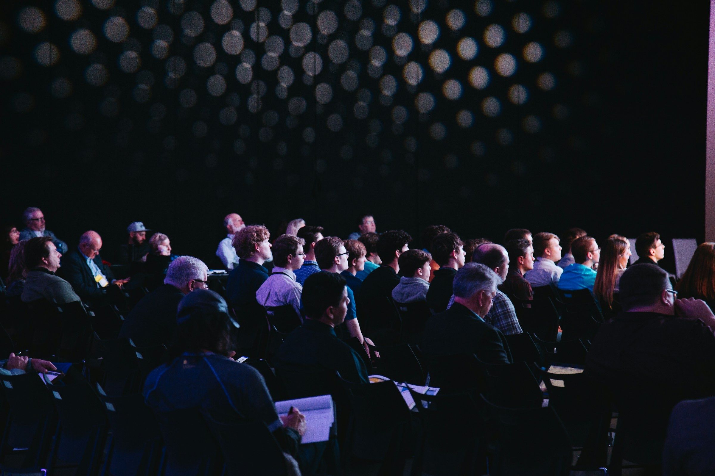 Audience members seen from behind watching a talk in a dark hall