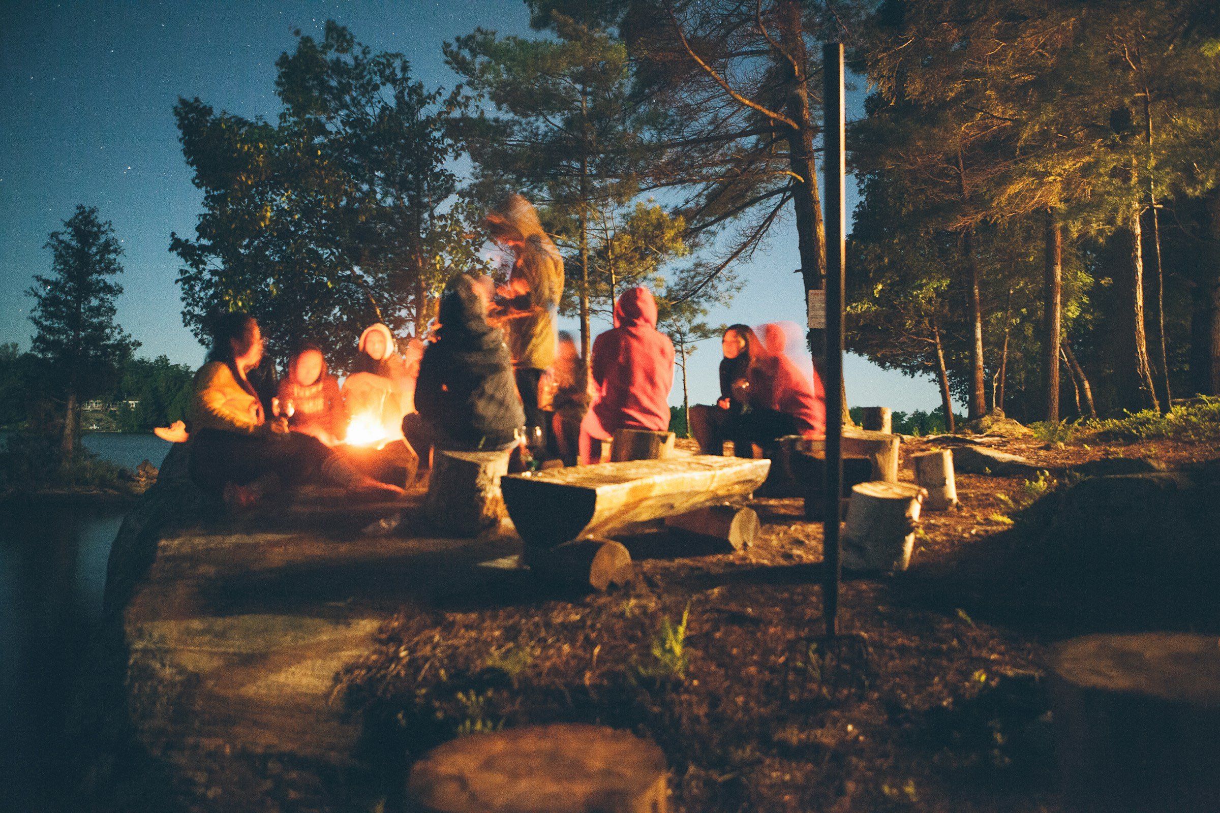 People around a campfire by a lake at dusk, seen from a distance