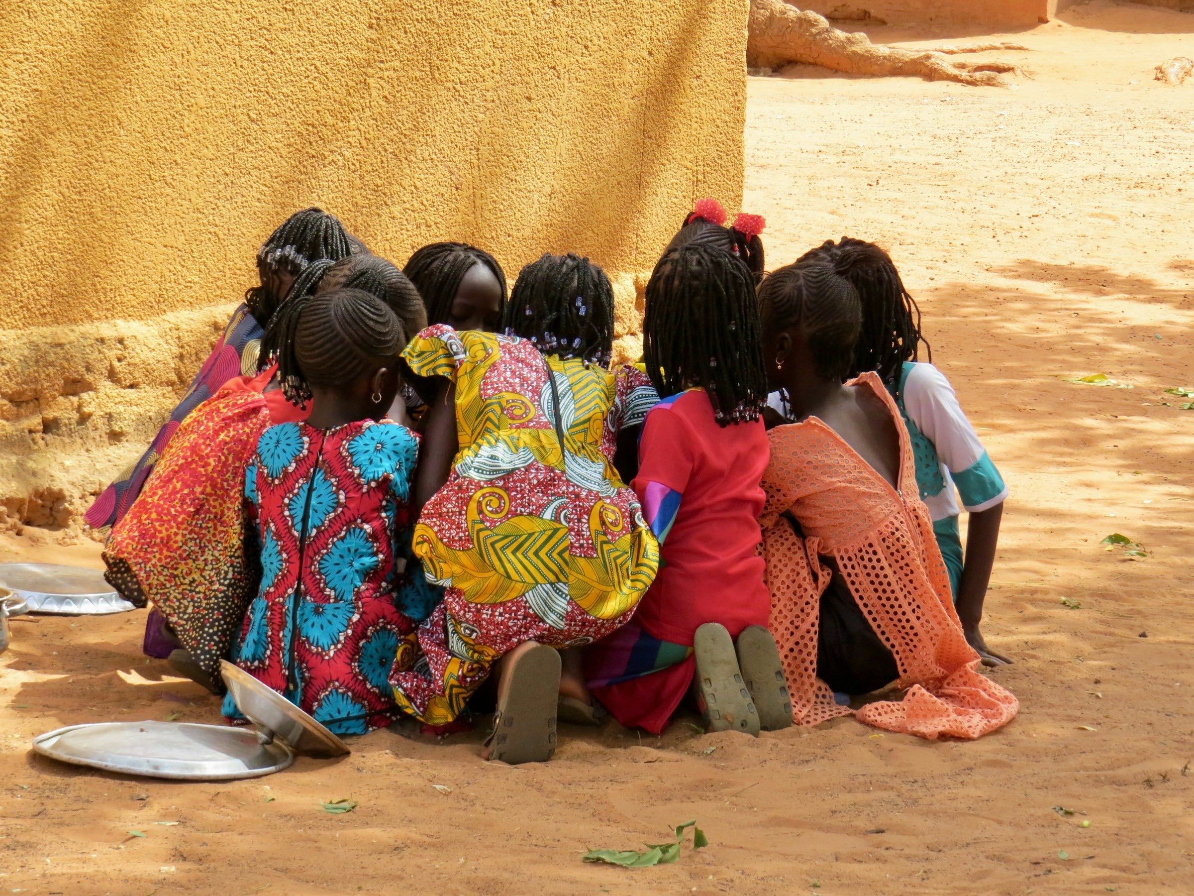 Children in colourful clothes crouched in a circle on red earth, seen from behind